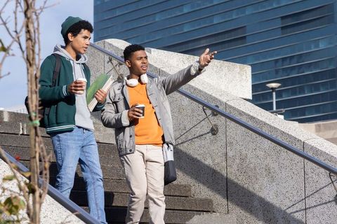 Diverse teen friends walking urban stairs holding coffee and notebooks, pointing ahead