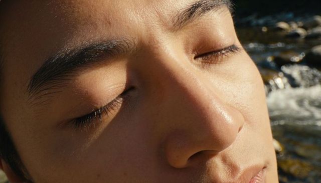 Basking asian male face soaking warm sunlight beside creek, closed eyes closeup serenity