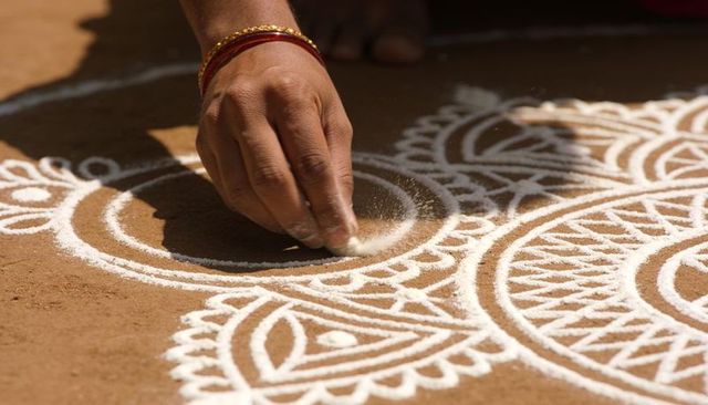 Woman decorating rangoli with white powder on clay courtyard, traditional indian craft