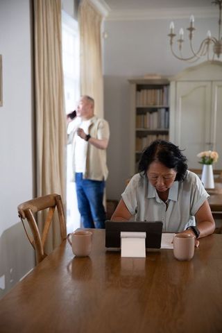 Senior chinese couple at dining table, woman using tablet while man talking on phone
