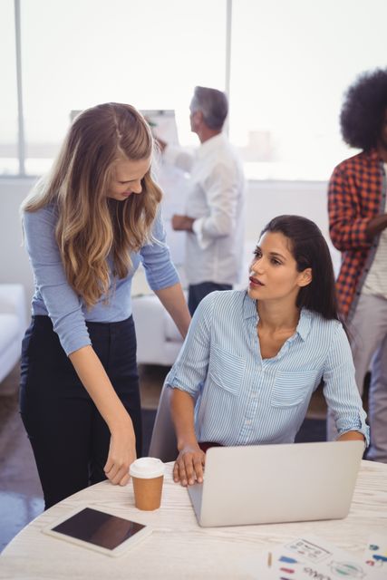 Two businesswomen are collaborating in a modern office. One is sitting at a desk with a laptop and coffee, while the other is standing and engaging in discussion. This image can be used to depict teamwork, professional collaboration, and a productive work environment in corporate settings.