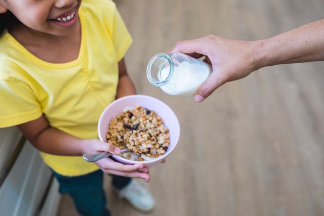 Cropped hand of hispanic man pouring milk in bowl held by smiling daughter at home. unaltered, healthy eating, family, lifestyle and togetherness concept.