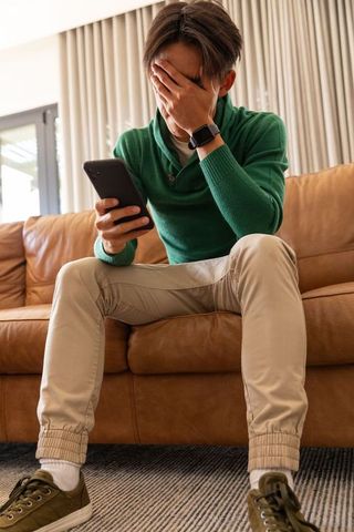 Young man covering face while reading smartphone on leather couch, feeling stressed