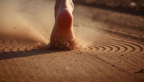 Barefoot heel pressing into golden sand creating airborne spray and curved raked grooves at sunset