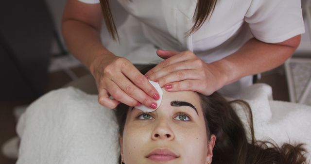 Image showing beautician applying eyebrow tint to woman lying down at beauty salon. Useful for content related to beauty treatments, professional services, spa visits, skincare routines, women’s self-care, or beauty industry promotions.