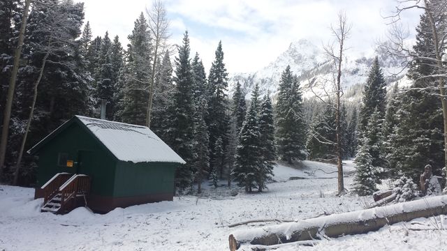 Snow covered cabin located in a remote mountain forest with snow-laden trees and distant mountain peaks in the background. Ideal for themes related to winter, nature, outdoor activities, holiday retreats, and serene getaways.