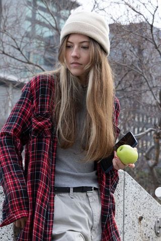 Young woman leaning on railing holding green apple and phone wearing beanie and plaid shirt