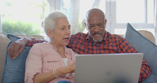 Senior couple sitting together on couch using laptop at home. Ideal for content promoting technology for seniors, retirement living, online activities for elderly individuals, or family bonding moments. Great for campaigns focusing on active aging, digital literacy among older demographics, and comfortable home living environments.