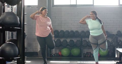 Diverse women balancing on one foot stretching in gym near dumbbell rack natural light