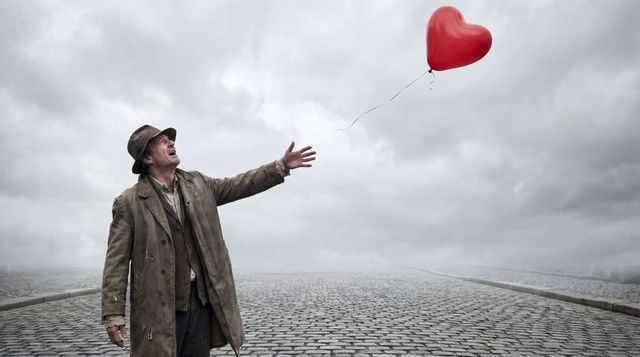 Solitary man reaching for red heart balloon on foggy cobblestone road, melancholic mood