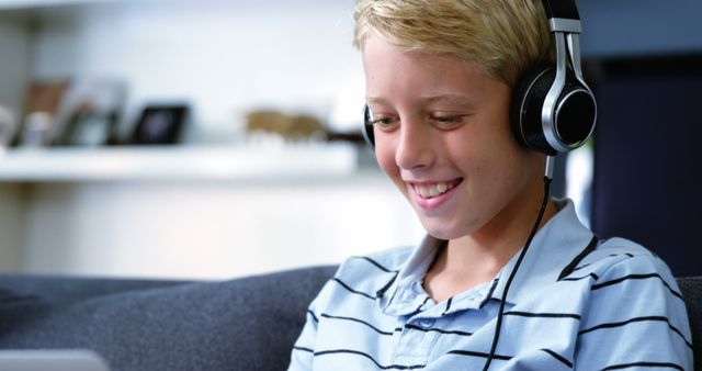 Boy is wearing headphones looking at laptop, suggesting he is engaged in virtual learning or an online class. His relaxed and happy expression conveys a positive educational experience. A useful image for illustrating remote education, e-learning platforms, educational technology, or children's use of gadgets at home.