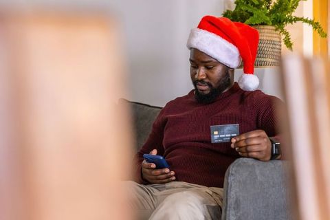 African american man wearing santa hat shopping online with credit card on couch