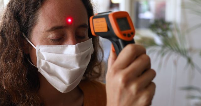 Caucasian woman spending time at home during coronavirus, covid19 lockdown, wearing a face mask, measuring temperature with a thermometer. Social distancing and self isolating.