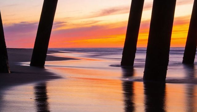 Pier pilings glowing at sunset over wet sand reflecting colorful dusk tides