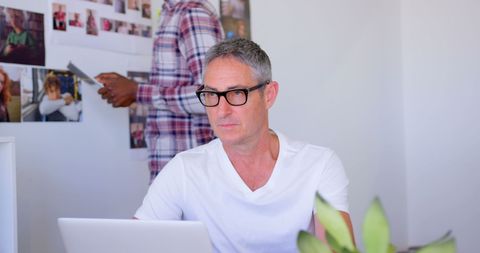 this visual depicts a determined mature male executive engaged in work on a laptop in a contemporary office environment. his attire is crisp and informal, signifying a relaxed yet productive atmosphere. in the background, a colleague of african descent is occupied with a digital tablet, showcasing a collaborative setting. this image is suitable for articles or materials relating to remote work, modern workplace dynamics, or executive management. it could enhance content promoting efficiency in corporate environments, leadership qualities, consulting firms, and technological advancements.