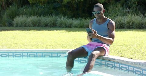 African American man relaxing poolside checking smartphone on sunny backyard lawn