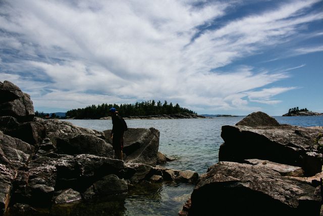 Person stands on rocky shoreline surrounded by water. The sky is partly cloudy, creating a dramatic backdrop. Islands and landmasses are visible on the horizon. Ideal for use in travel websites, adventure blogs, outdoor and hiking promotions, and nature-themed content.