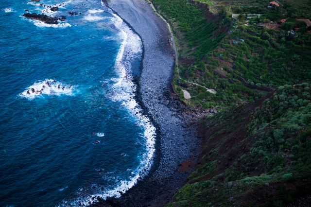 Aerial view of a dramatic rocky coastline with waves rolling onto the shore next to oceanfront cliffs. The blue water contrasts with the dark rocks and lush green foliage on the cliff. Perfect for travel blogs, nature magazines, and environmental campaigns.