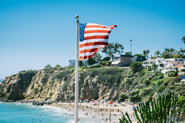 American flag waving on flagpole overlooking a sunny California beach packed with people enjoying the summer. Cliffside homes and lush nature add to the scenic view, making it perfect for themes related to patriotism, travel, summer vacations, and outdoor activities.