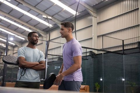 Diverse male friends chatting beside indoor padel court holding paddles, casual training
