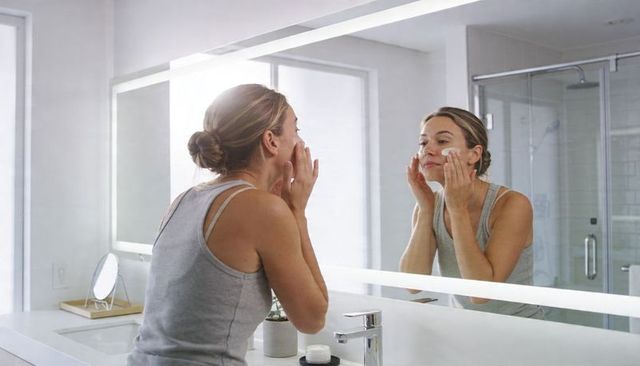 Woman applying moisturizer in bright minimalist bathroom mirror during daily skincare routine