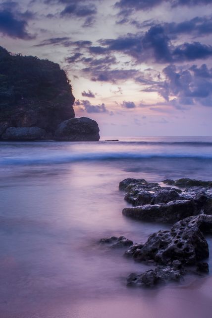 This stunning beach scene captures the tranquility of a tropical coastline during sunset. With gentle waves lapping against rocky shores and a vibrant purple sky, it exudes calmness and natural beauty. Perfect for travel websites, nature magazines, or wall art centered around peaceful seaside themes.