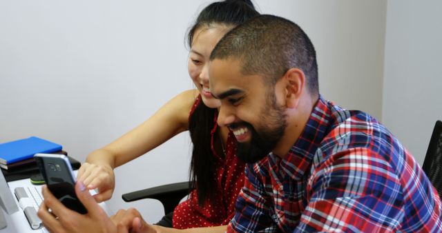 Young man and woman coworkers sitting at desk, smiling and using smartphone in an office setting. Ideal for themes of teamwork, modern office environment, technology use in the workplace, and positive coworker interactions.