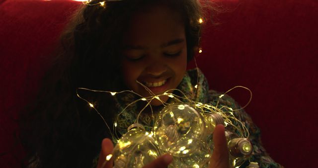 Young girl enjoying holiday season, holding glowing fairy lights with joyful expression. Ideal for use in holiday greeting cards, festive advertisements, children's magazines, and promotional materials celebrating the magical season.