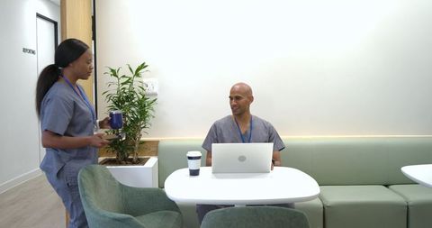 African american nurses in gray scrubs chatting in staff lounge with laptop and coffee