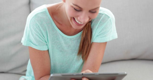 Young woman using a digital tablet while sitting on a couch. Ideal for use in lifestyle blogs, technology advertisements, and home living articles. The relaxed and happy expression suggests themes of enjoyment and ease when using digital devices at home.