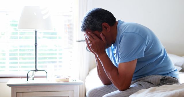 Man is seen covering face while sitting on bed, capturing a moment of stress or worry. Indoor scene with soft lighting emphasizes emotional impact. Can be used in mental health awareness campaigns, articles about stress, or images illustrating emotions such as sadness or concern.
