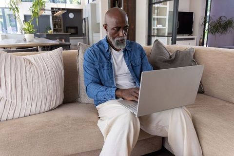 Senior african american man working on laptop on beige couch in modern open-plan living room