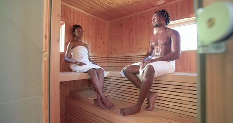 African couple relaxing in wooden sauna wrapped in white towels for spa wellness retreat