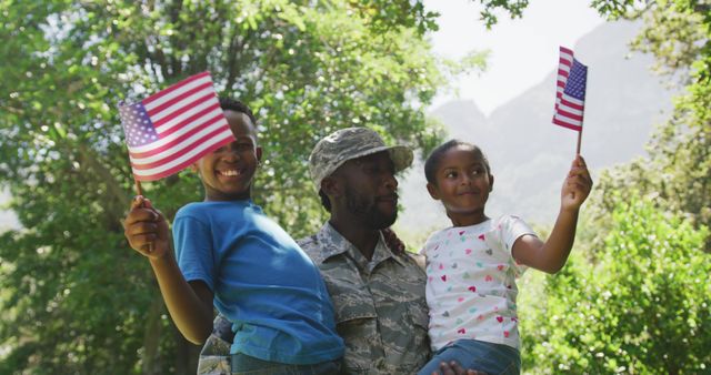 This image shows an African American soldier embracing his children who are holding American flags. They are outdoors surrounded by lush greenery and trees, creating a bright and natural background. Perfect for themes related to patriotism, family bonding, military appreciation, and national holidays like Independence Day or Memorial Day.