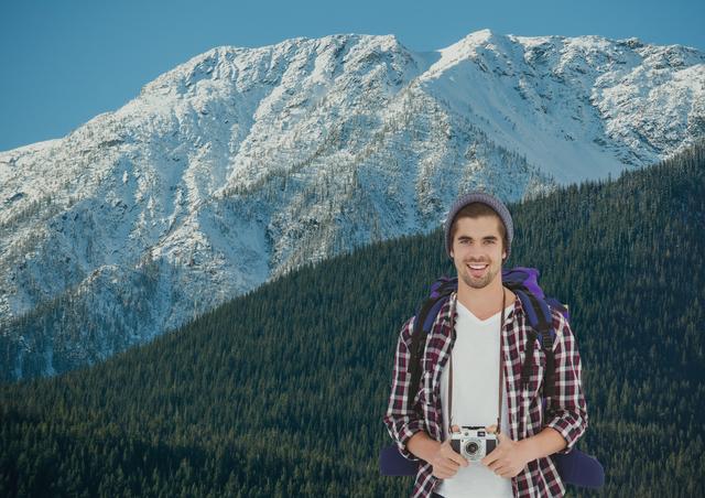 A young man wearing a beanie, holding a camera, and smiling during a hike. Snow-covered mountain in the background. Ideal for travel blogs, adventure magazines, tourism campaigns, and outdoor apparel promotions.
