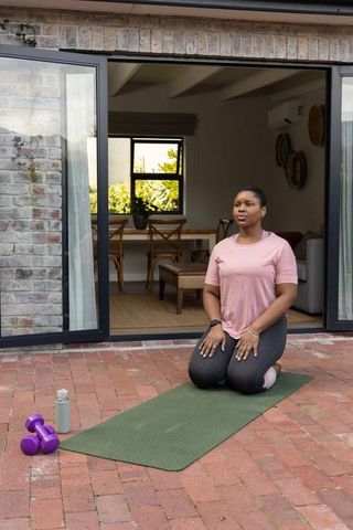 African american woman practicing outdoor home workout kneeling on mat in courtyard with dumbbells