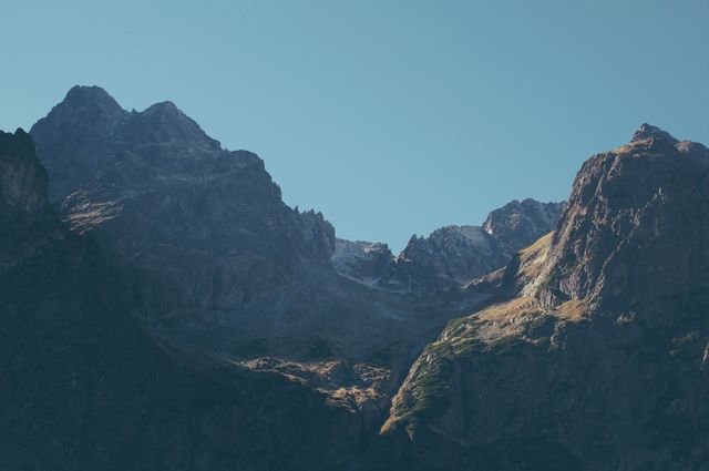 Dramatic sunlit mountain peaks with rugged rock formations under blue sky. Ideal for nature-themed website headers, travel blogs, adventure promotions, and outdoor activity marketing. Conveys tranquility, natural beauty, and exploration.