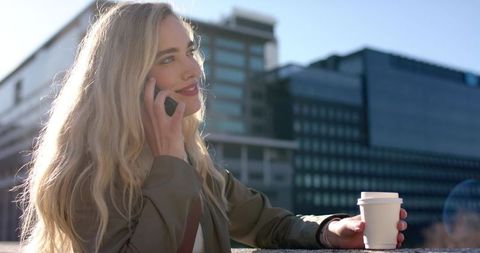 Young woman talking on phone while holding coffee on urban balcony during golden hour