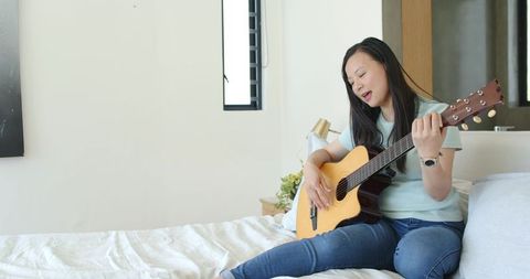 Asian woman playing acoustic guitar on bed singing in cozy daylight bedroom