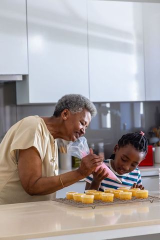 Grandmother guiding granddaughter piping pink icing onto cupcakes in modern kitchen