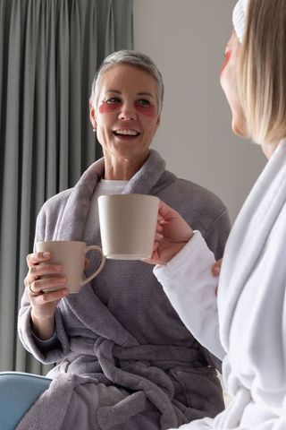 Mature women sharing morning coffee in plush robes, wearing under-eye gel patches, relaxing