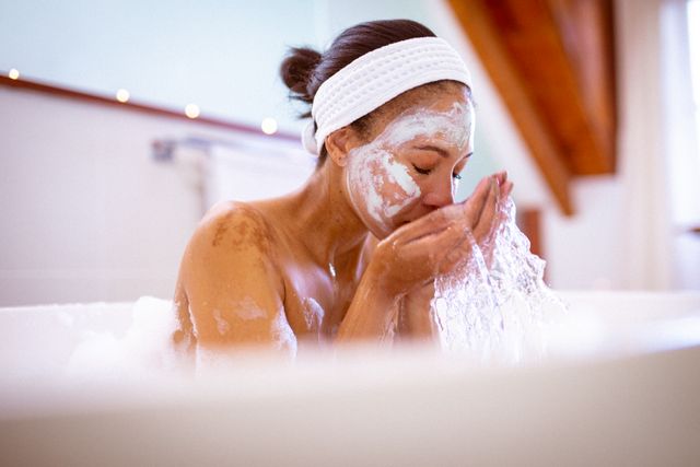 Biracial woman enjoying a relaxing bath while cleaning her face. Perfect for promoting self care, wellness, and beauty treatments. Ideal for use in articles, blogs, and advertisements related to personal hygiene, home spa experiences, and domestic lifestyle.
