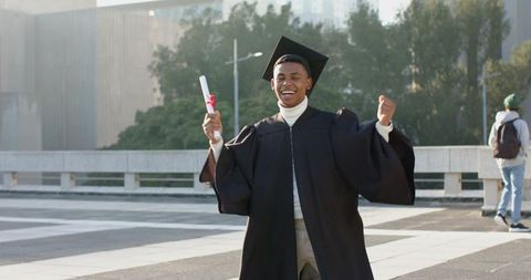 Joyful african american graduate celebrating with diploma in cap and gown on campus plaza