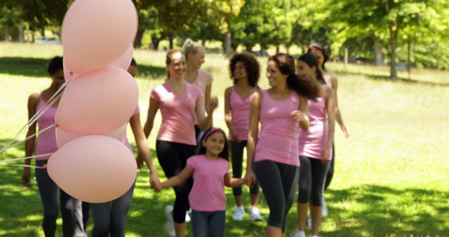 Group of women and a young girl walking together in a park with pink balloons. They wear matching pink shirts, suggesting participation in an event like a walk for breast cancer awareness. Useful for promoting community events, health and wellness campaigns, and breast cancer awareness activities.