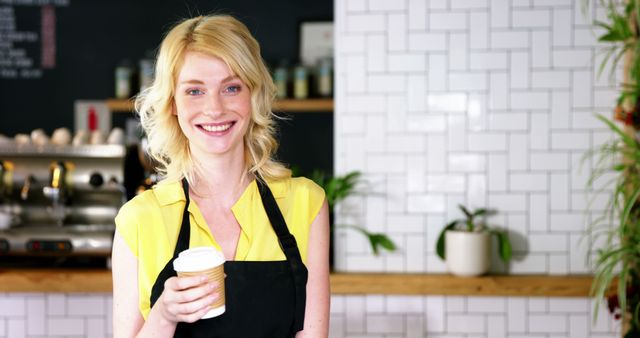 Blonde female barista smiling while holding a coffee cup in a modern cafe. Perfect for concepts related to customer service, coffee culture, hospitality industry, and small business promotions.