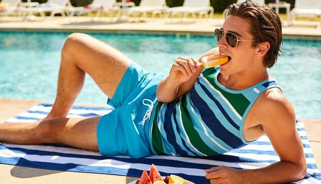 Young man relaxing poolside, eating orange popsicle and sunbathing on striped towel
