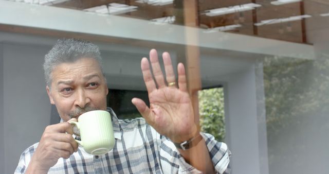 Elderly man enjoying a cup of coffee while waving to someone through a window in a comfortable home setting. Ideal for use in marketing materials related to lifestyle, senior living, morning routines, and family connections.