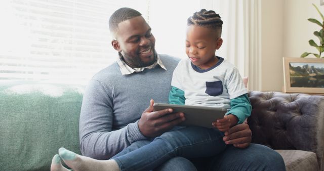 Father and son bonding while using a tablet together in a cozy home environment. They look engaged and happy, sitting closely and sharing a moment. Ideal for use in advertising family-oriented services, parenting blogs, digital learning content, and technology use in everyday life.