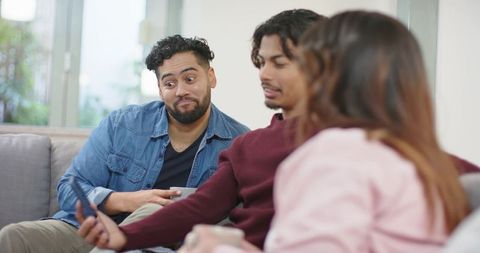 Diverse friends relaxing on sofa sharing smartphone and coffee in bright modern living room