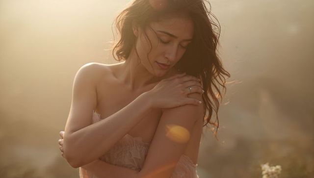 Backlit woman standing wearing strapless lace dress showing engagement ring at golden hour